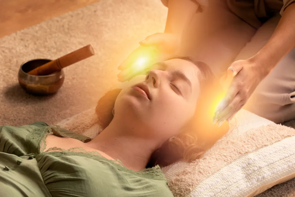 A practitioner holding glowing hands above a reclining client beside a singing bowl.
